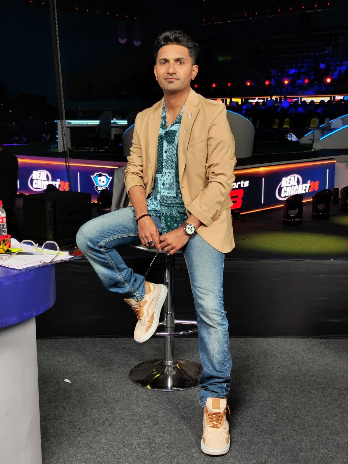 GEPL caster desk — beige blazer, denim, perched on the host stool with the Real Cricket 24 set behind